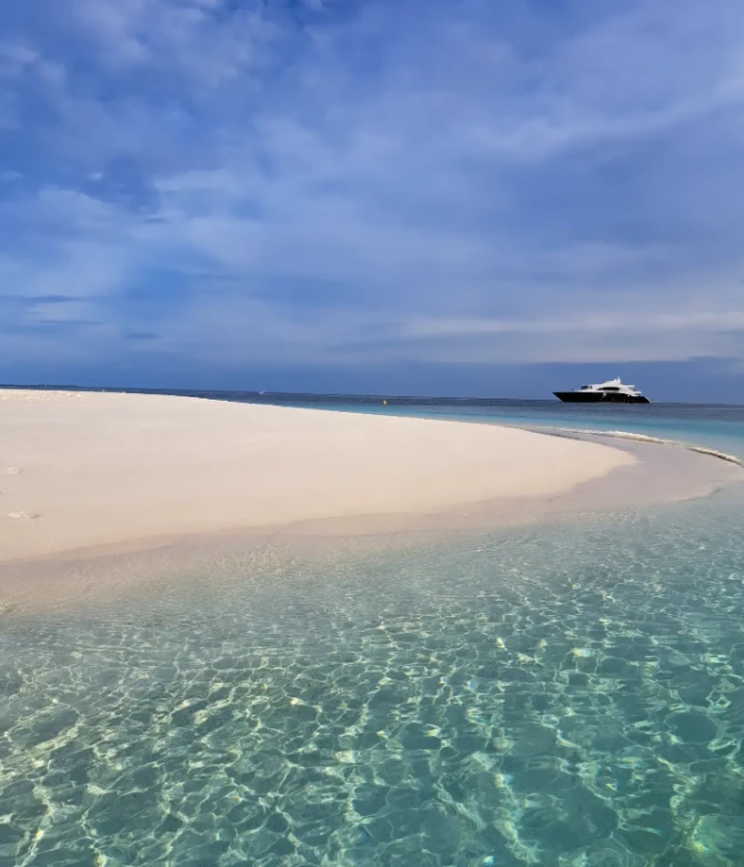 Clear turquoise water around a white sandbank, with a liveaboard anchored offshore