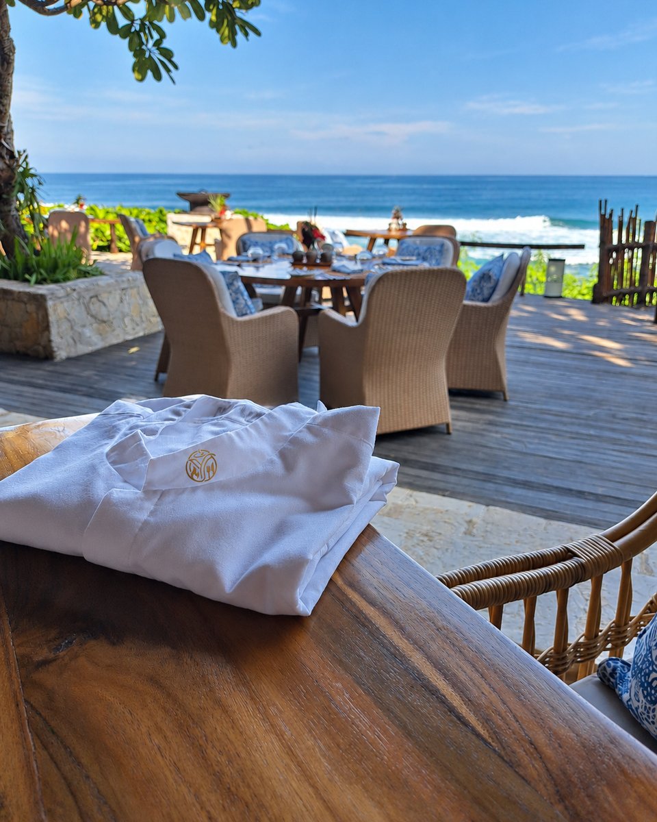A folded chef's jacket with the Yunic monogram on a wood table, a beachfront dining deck and the ocean beyond
